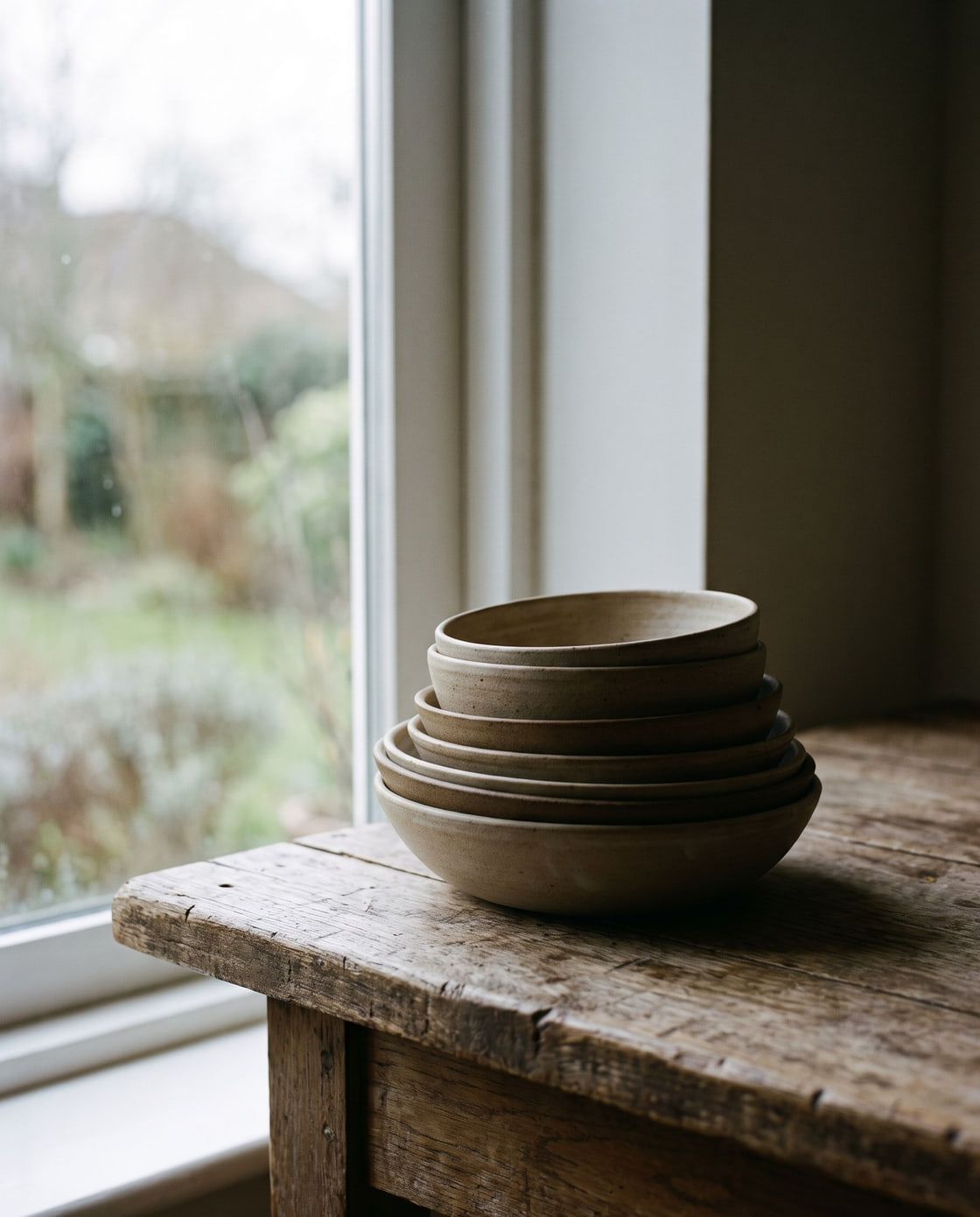 Stos nieszkliwionych ręcznie robionych misek stoi na surowym drewnianym stole przy oknie w miękkim świetle.