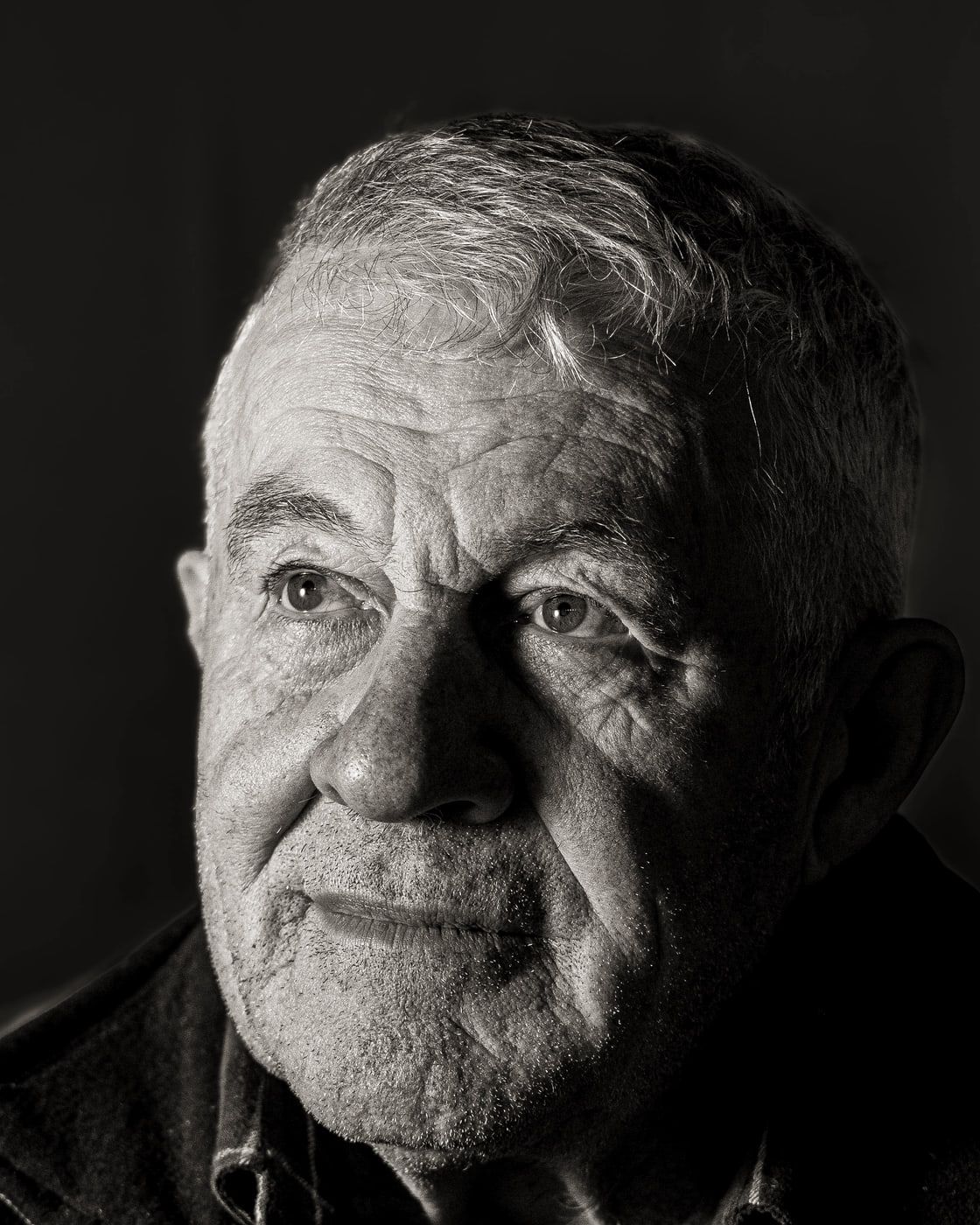 Czarno-biały portret starszego mężczyzny o zniszczonej twarzy i krótkich siwych włosach na ciemnym tle.